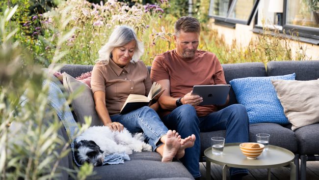 Ægtepar sidder i sofa ude på deres terrasse med deres hund. Kvinden læser en bog imens manden sidder på sin tablet og kigger på energiløsninger fra Andel Energi.